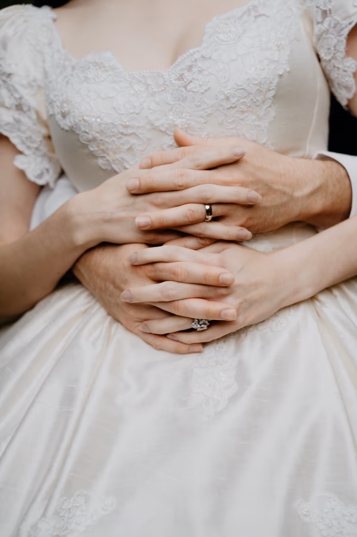Close-up of a bride and groom's hands intertwined on the bride's lace wedding dress, showing their wedding rings.