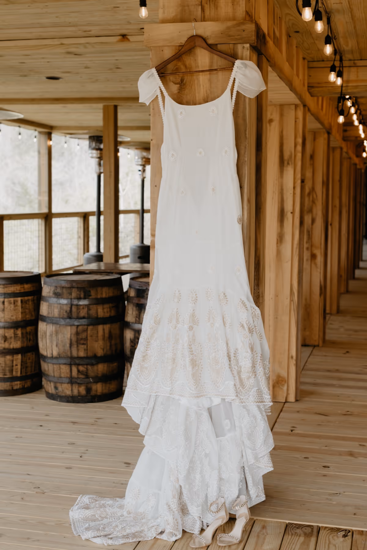 White wedding dress with lace details hanging on wooden wall with matching high-heeled shoes below on wooden floor.