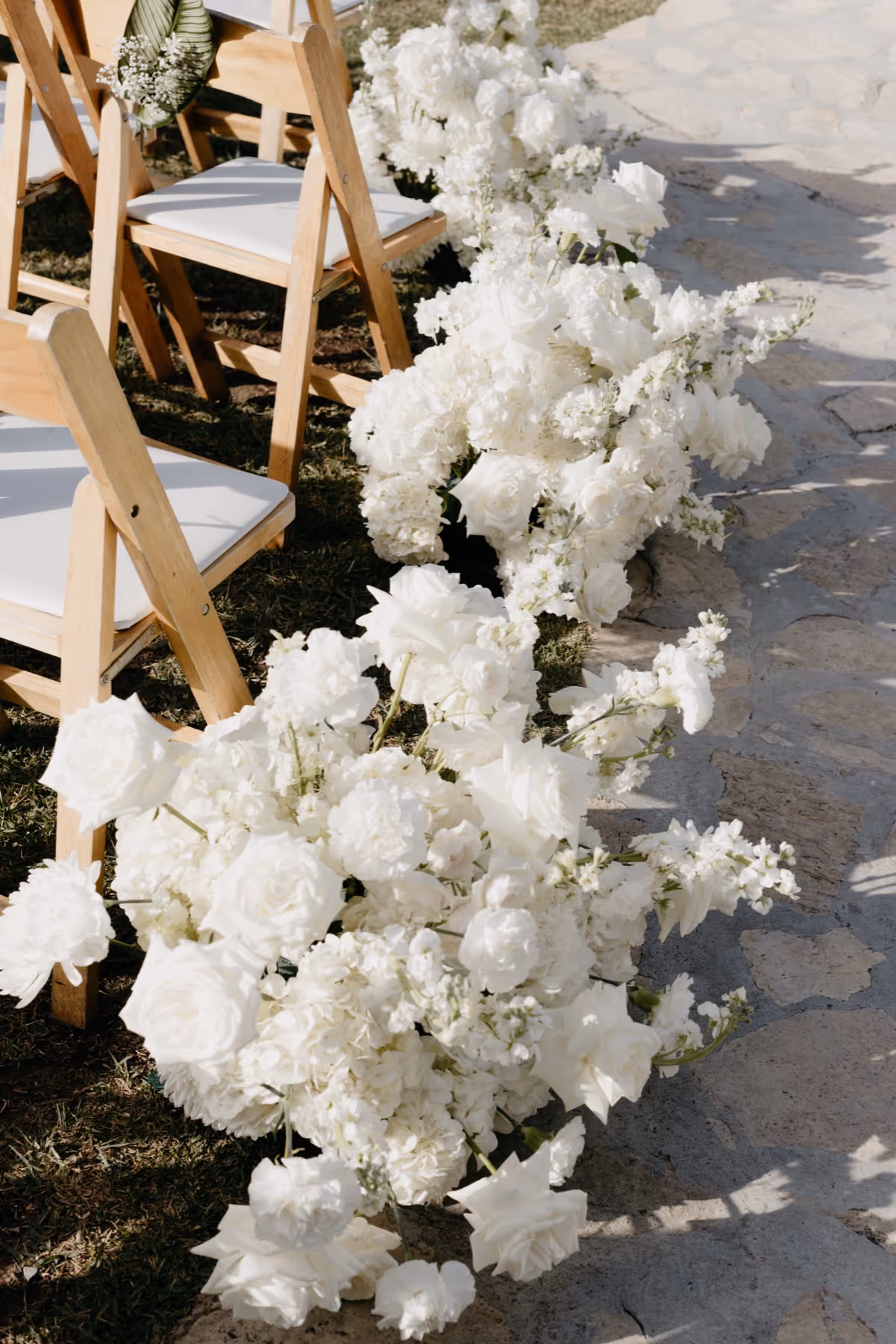Rows of wooden chairs with white cushions next to a stone pathway lined with large white floral arrangements.