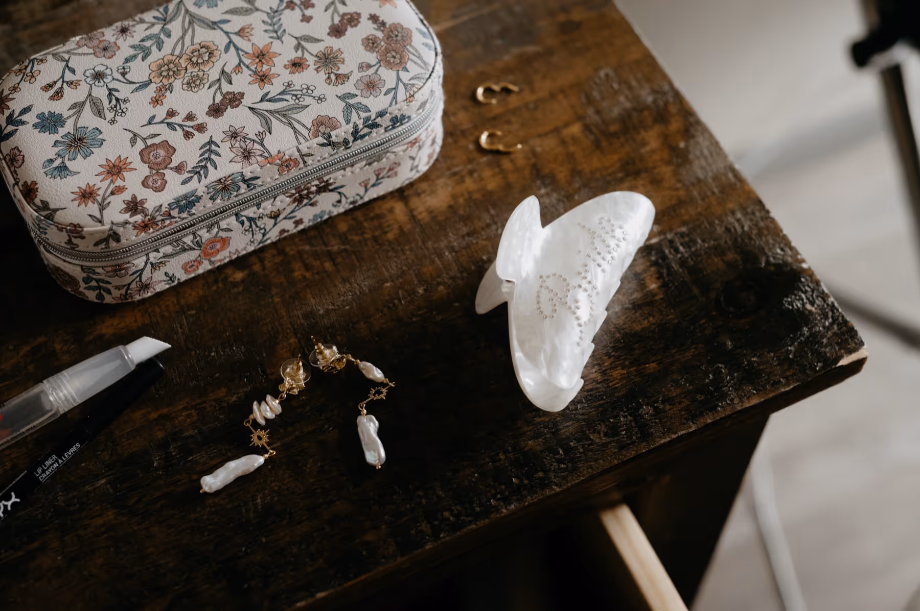 Floral-patterned makeup bag, pearl drop earrings, lip liner, and white hair clip on a dark wooden table.