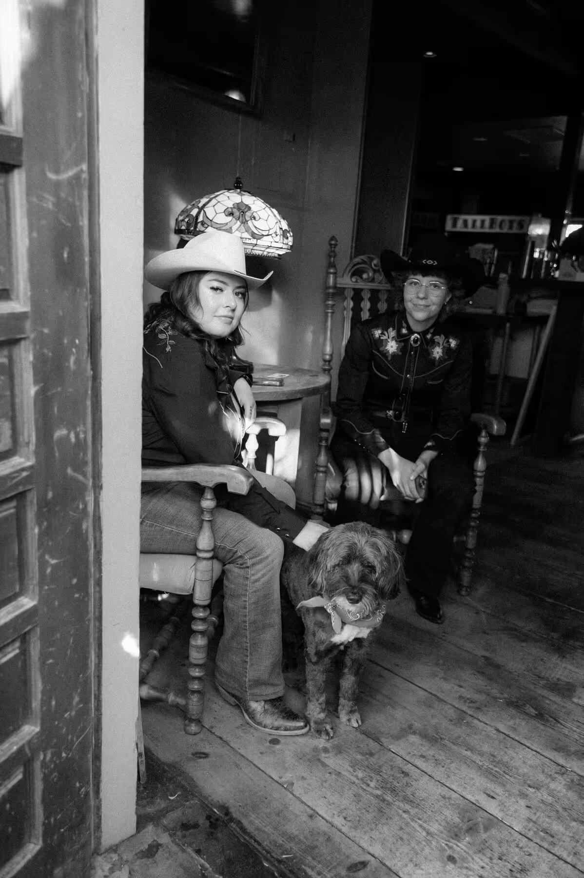 Two people in cowboy hats sitting on wooden chairs indoors with a dog standing between them on a wooden floor.
