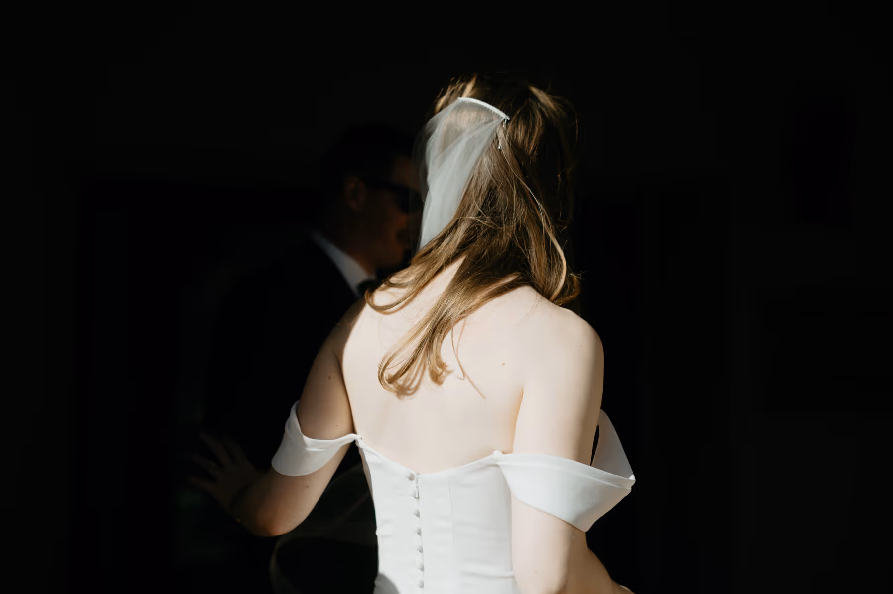 Bride in an off-shoulder white wedding dress with a veil, facing away, with a groom in the background.