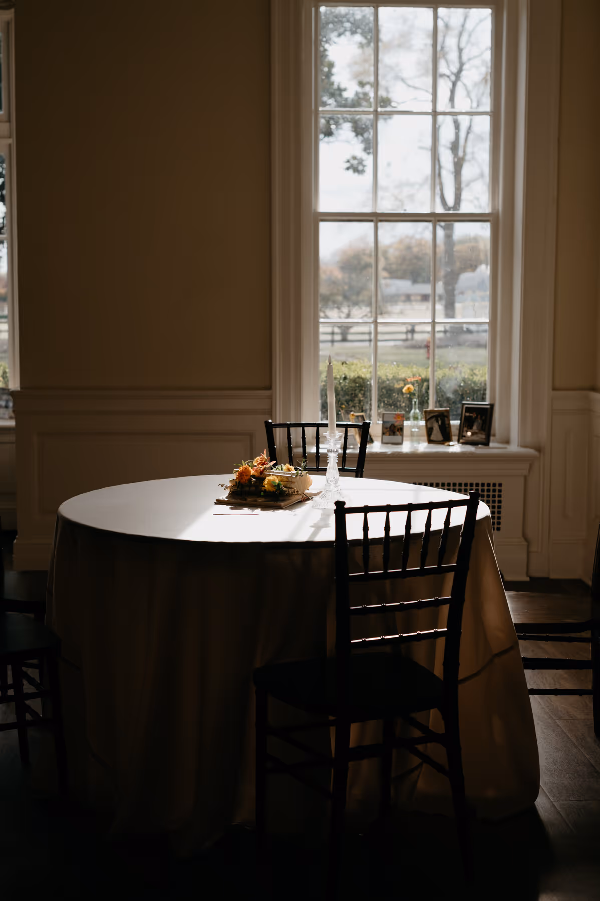 Round table with white tablecloth and floral centerpiece with a white candle, lit by sunlight near a large window.