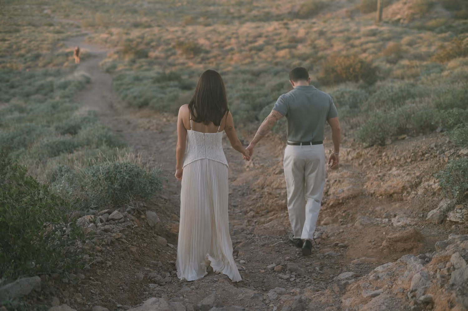 Couple holding hands walking down a rocky dirt path in a desert landscape with green shrubs.