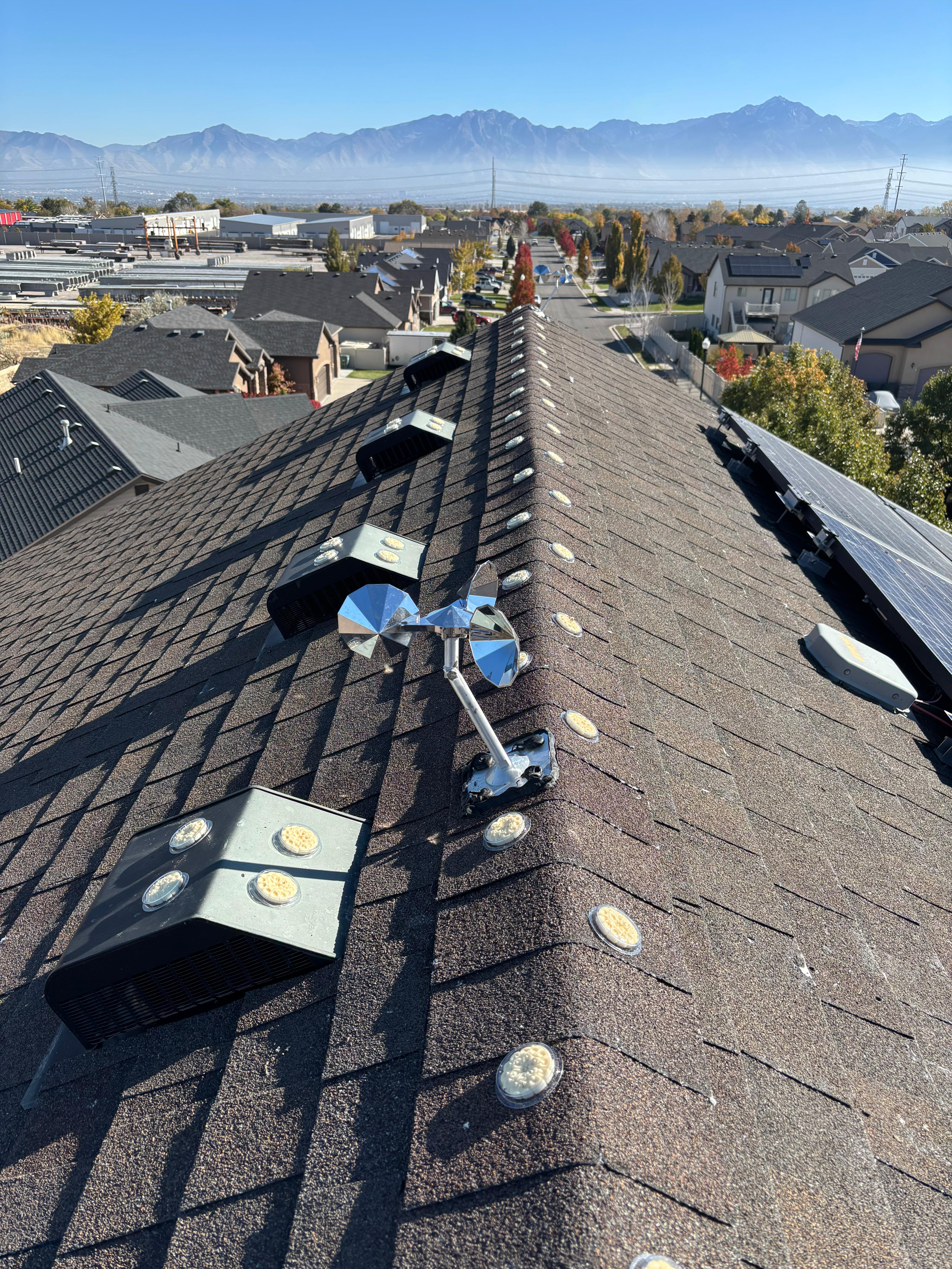 Shingled rooftop with Optical Gel, snow guards, a metal wind direction vane, vents, solar panels, and a suburban neighborhood with mountains in the background under a clear blue sky.