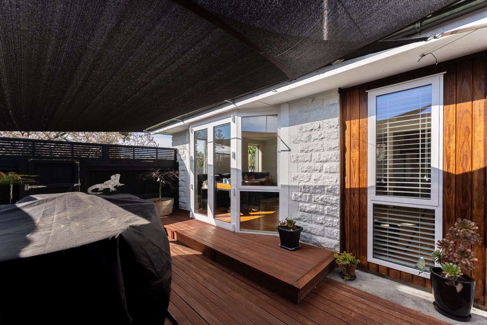 Outdoor wooden deck with a black canopy overhead, a covered grill on the left, potted plants on the right, and white-framed glass double doors and a window leading inside a house with white brick and wooden siding.