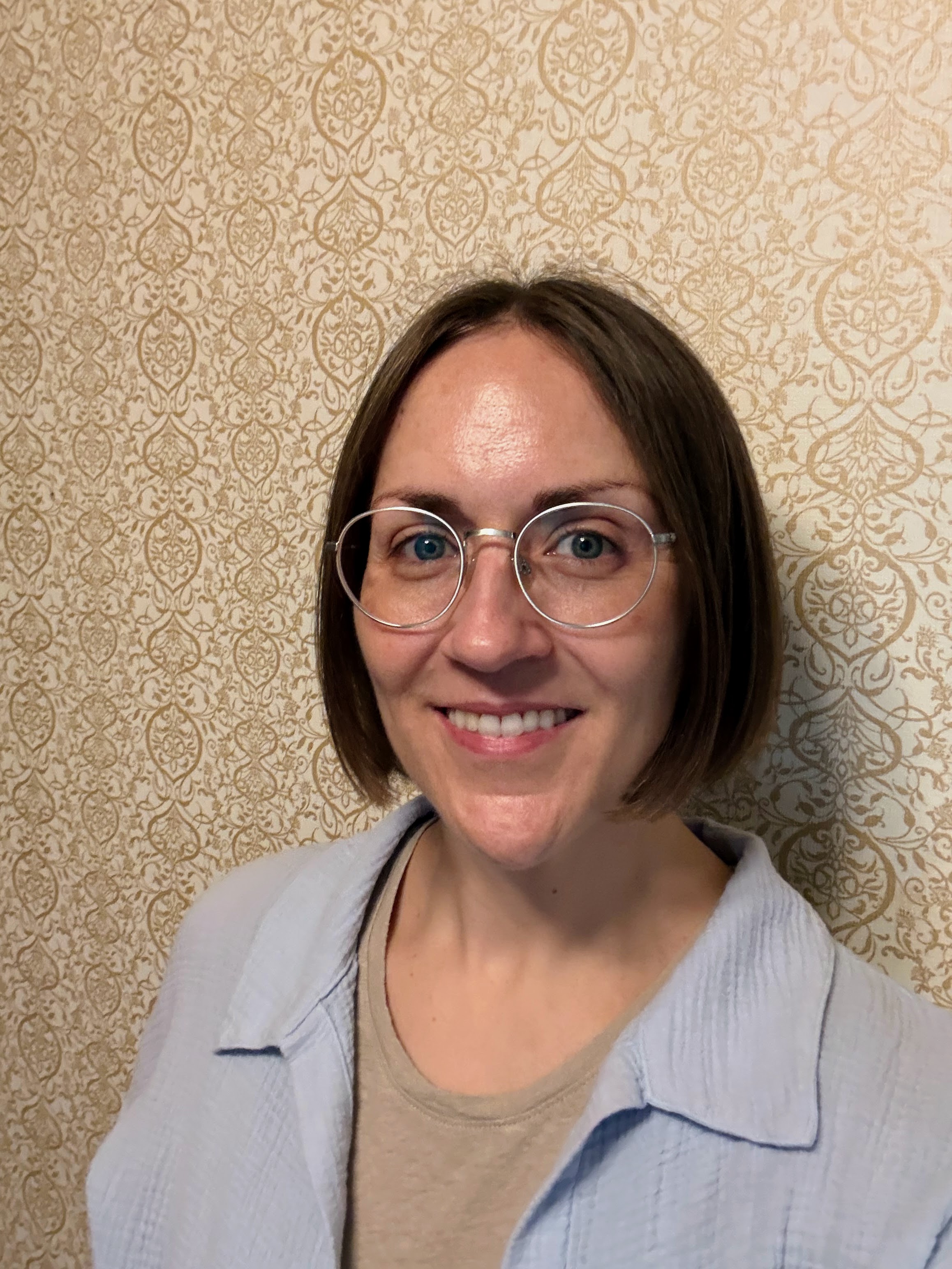 Testimonial provider. Smiling woman with short brown hair wearing round clear glasses, beige shirt, and light blue jacket, standing in front of patterned wallpaper.
