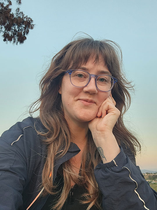Testimonial provider. Smiling woman with short brown hair wearing round clear glasses, beige shirt, and light blue jacket, standing in front of patterned wallpaper.