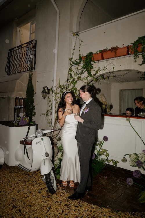 Bride in a white dress and groom in a dark suit holding drinks and smiling at each other beside a white Vespa bar outside a building with plants.