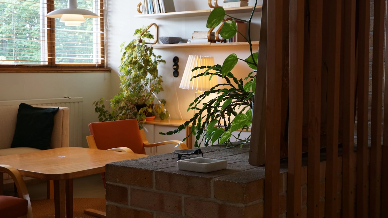 Cozy office corner with wooden table, chairs, green plants, a lamp, and window blinds.