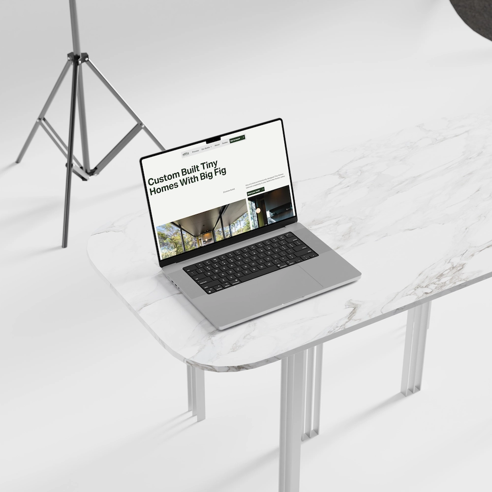 Open laptop on white marble table displaying a webpage titled 'Custom Built Tiny Homes With Big Fig' with images of homes, set in a minimal white studio with a tripod light stand in the background.