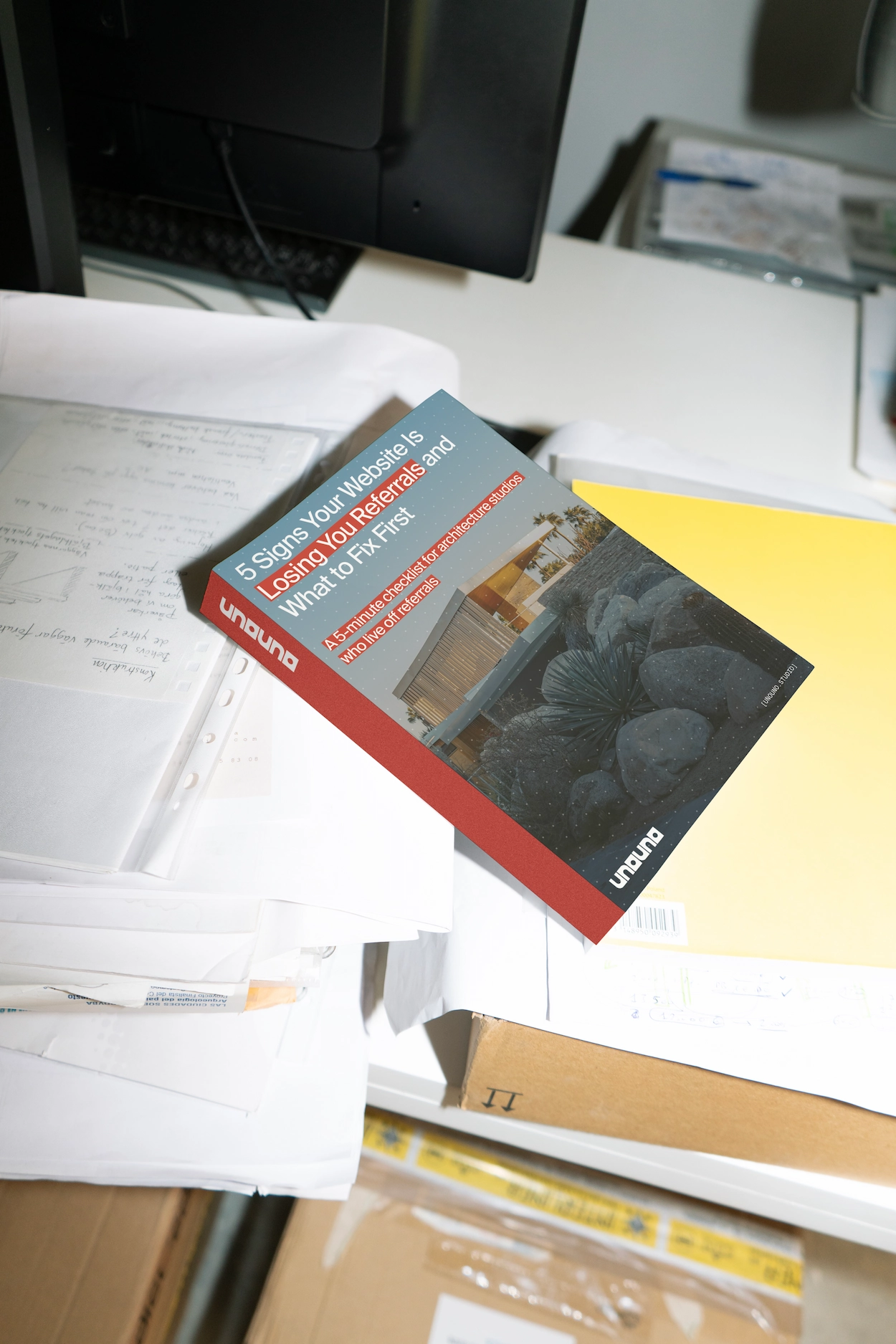 A book or booklet titled '5 Signs Your Website Is Losing You Referrals and What to Fix First' with a subtitle about a 5-minute checklist for architecture studios who live off referrals, resting on a cluttered white desk with papers, a yellow folder, and a computer monitor in the background.