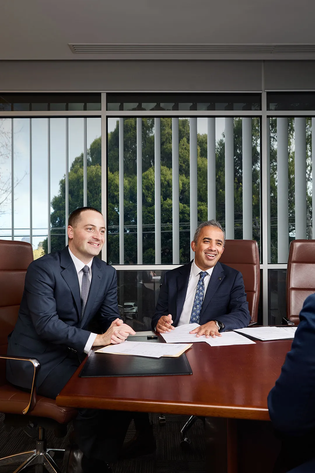 Two businessmen in suits sitting and smiling across a wooden conference table with documents, in a bright office with large windows and vertical blinds.