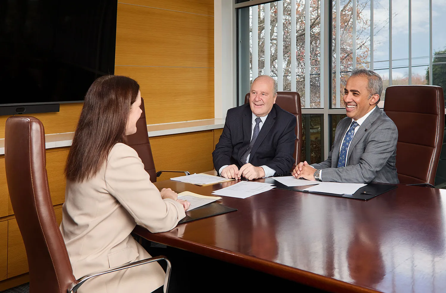 Two men in suits smiling and talking with a woman in a beige blazer during a meeting at a conference table with documents.
