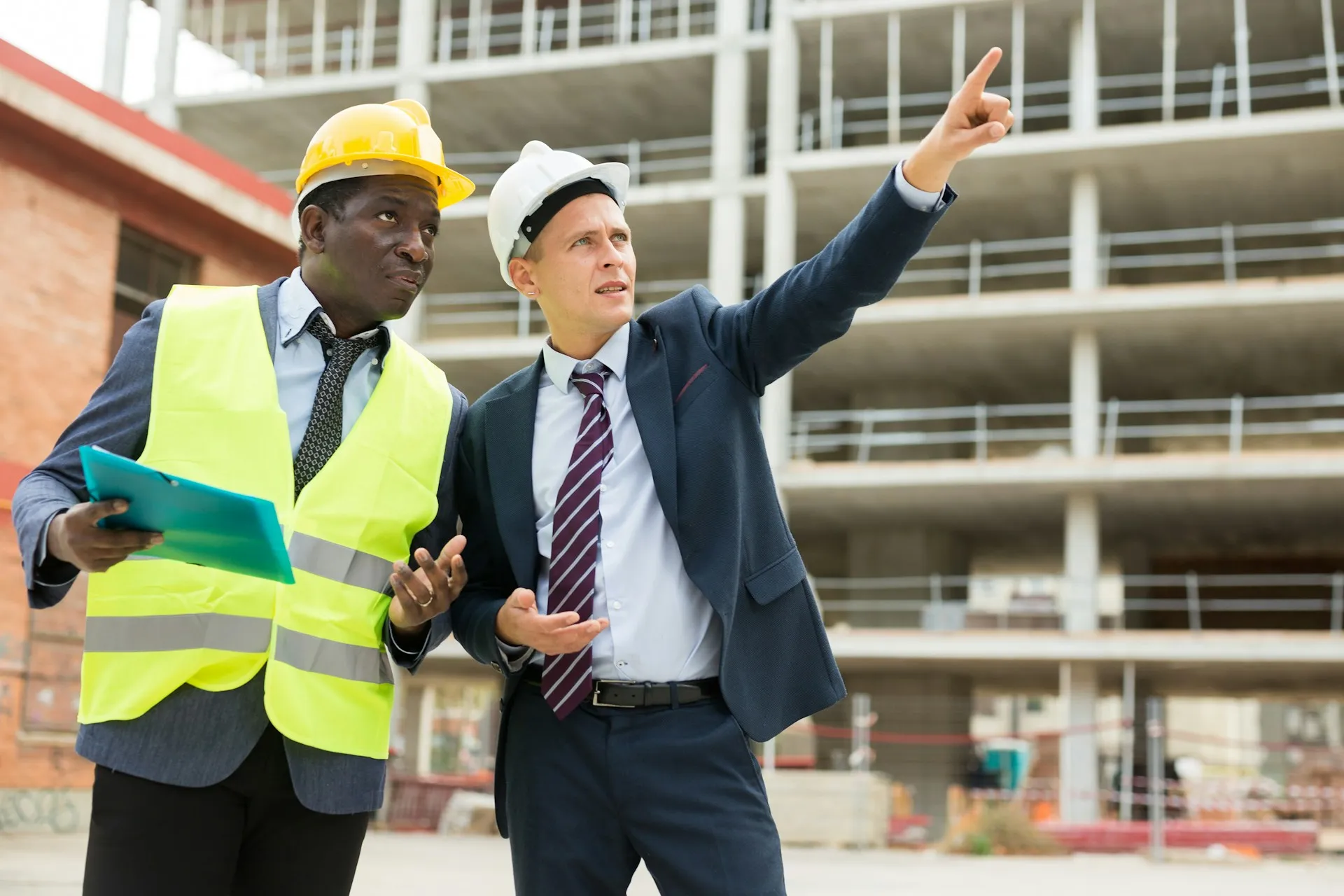 Two construction professionals wearing hard hats and formal attire discuss plans at a building construction site, with one pointing forward.