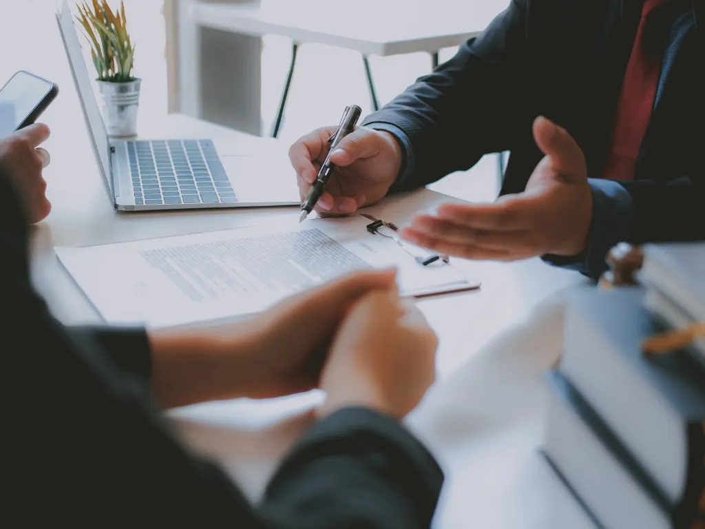 Two people in a business meeting reviewing a document on a clipboard with a laptop and a small plant on the table.