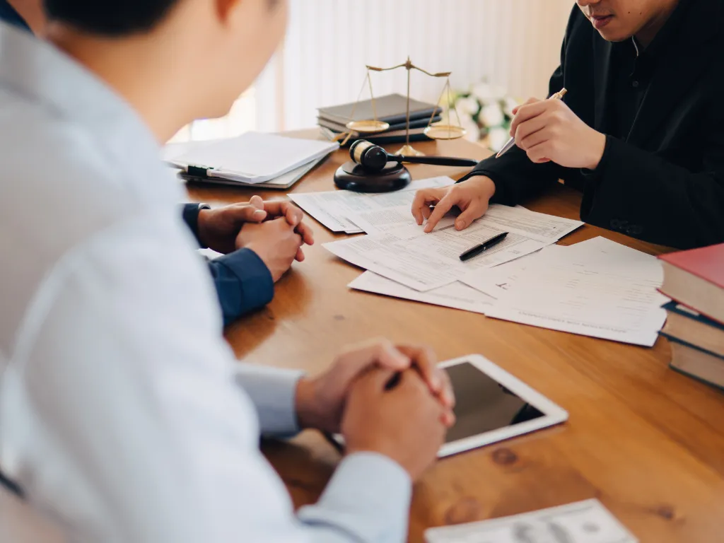 People discussing legal documents at a wooden table with a gavel, scales of justice, and books in the background.
