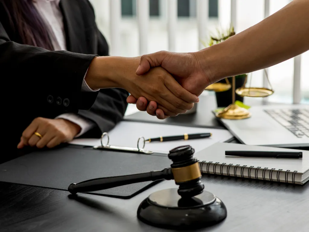 Two people shaking hands over a desk with a judge's gavel, legal scales, and documents, symbolizing a legal agreement.