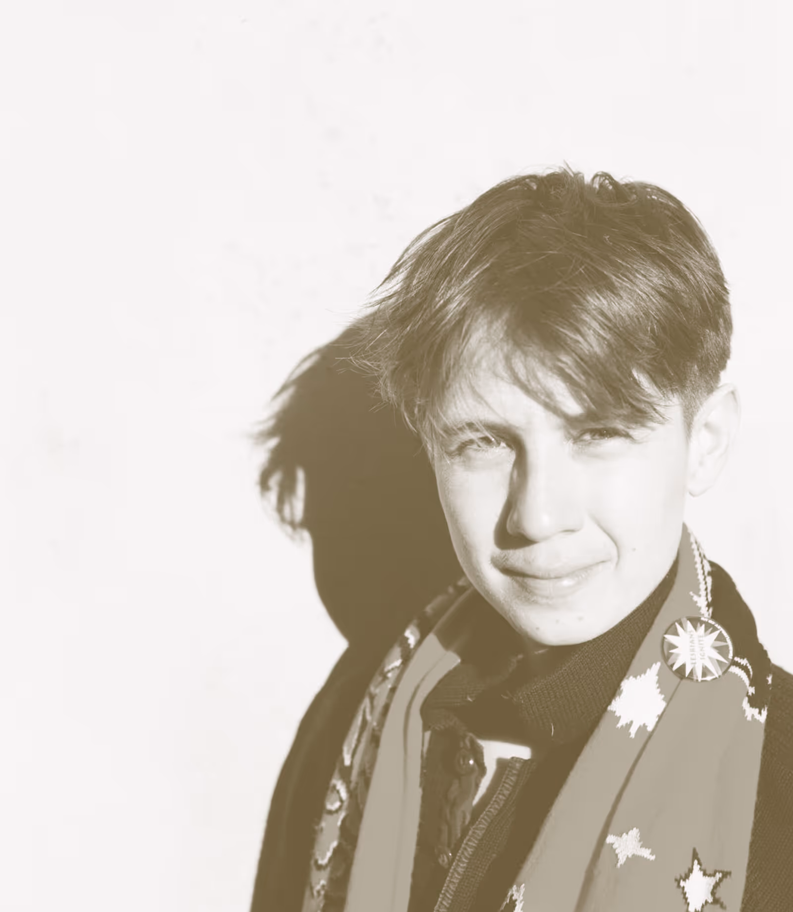 A portrait image with a goldy brown filter, of a person from the shoulders up facing the camera and smiling. They have short hair that flops across their eyes and wearing a scarf and button up jacket.