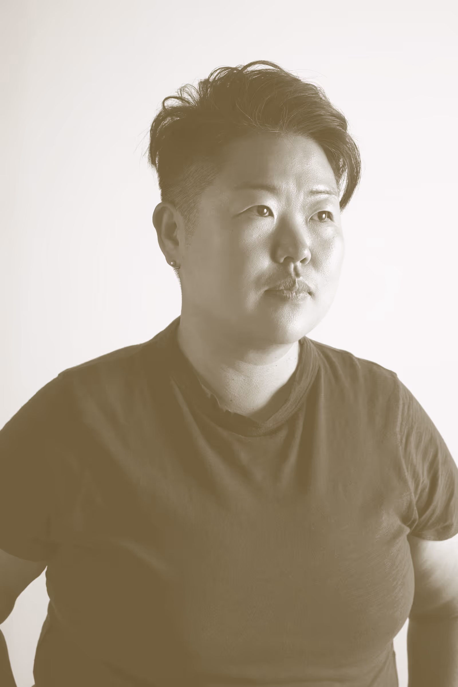 A portrait image with a goldy brown filter, of a person from the shoulders up facing stage left with a neutral expression. They wear a plain t-shirt and studs in their ears and have a side part cut.