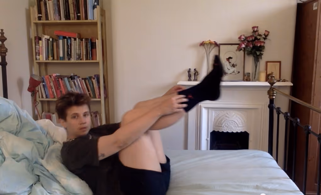 A landscape image of a slightly blurred figure on an iron post bed wearing a black t-shirt, shorts and socks. The figure is falling onto their back with their legs bent in the air, arms stretched out in front of them pulling their socks up. Behind them, bundled up are the bedclothes. The figure is looking at the camera slightly surprised. In the background a full bookcase fills the left corner of the image, a sliver of a wooden wardrobe the right corner. Towards the middle right is an old fireplace and mantle, painted white like the walls. On the mantle are figurines, pictures in frames, bunches of flowers and candles.