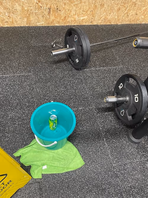 A portrait image. The ends of two black 10kg and 15 kg weights on steel bars enter the image from the right, they are resting on a speckled tiled floor and labelled with the Pure Gym symbol. Next to them on the floor to the left is a turquoise bucket sitting on top of a slightly scuffed up lime green microfibre cloth. There is a half crushed can of Boost in the bucket and the corner of a yellow ‘wet floor’ sign in the lower left corner of the image.