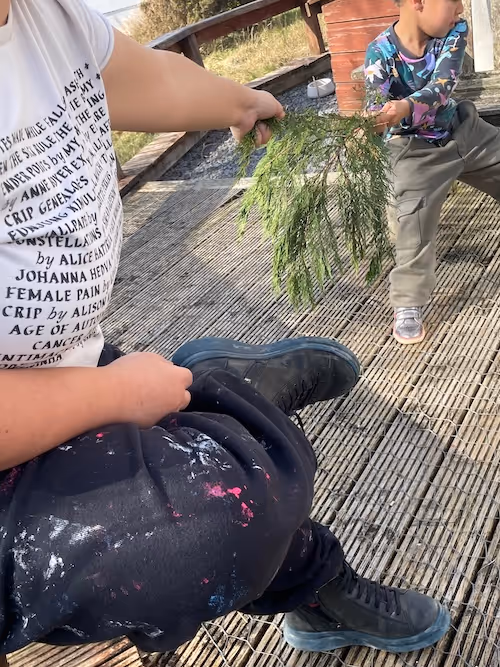 A portrait image of the front of a figure leaning into the picture. The front of a white t-shirt with black serif cursive writing out (indistinguishable) names, black paint spattered trousers and black and blue trainers. The figure  reaches towards a child in the upper right corner of the picture wearing a patterned top, grey trousers and silver trainers, extending out a branch of greenery. Both figures sit on a platform of wooden decking with grasses and flinted slate in the background.