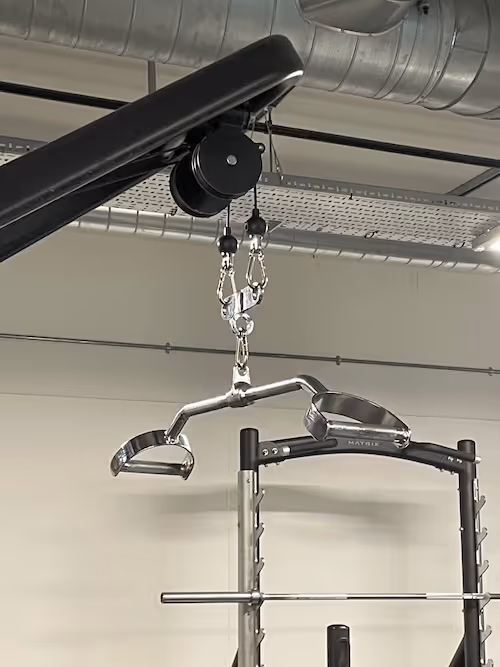 A portrait image, at the forefront a steel pulley system with a bar and two hand placements that resemble the shapes of stirrups or the handle of spades, hangs from a black overhang. In the background a black and steel frame for a weight bench with a steel bar sits to the right side of the image. Cylindrical silver air vent systems fill the upper part of the background.