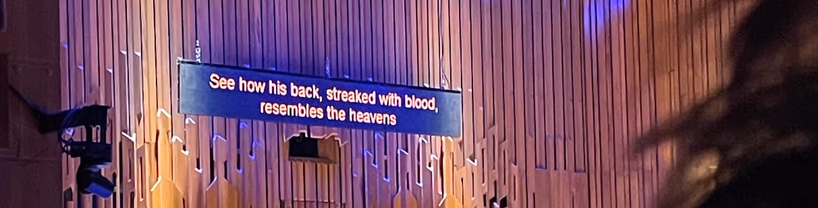 A thin cropped landscape image of the top of a stage set built of wooden slats to form an intricate pattern. The right corner is blurred by a shadow of a figure, the left shows the beginning of stage lighting. In the middle a stage captioning monitor is suspended with the following text in digital orange: “See how his back, streaked with blood, resembles the heavens”.