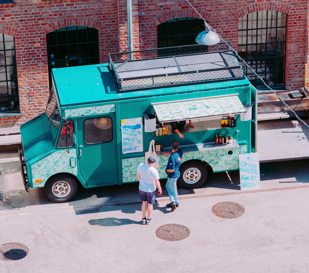 En turkis matvogn står parkert i en gate ved en murbygning. To personer står ved serveringsvinduet, den ene bestiller og den andre venter. Sollyset kaster skygger på bakken.