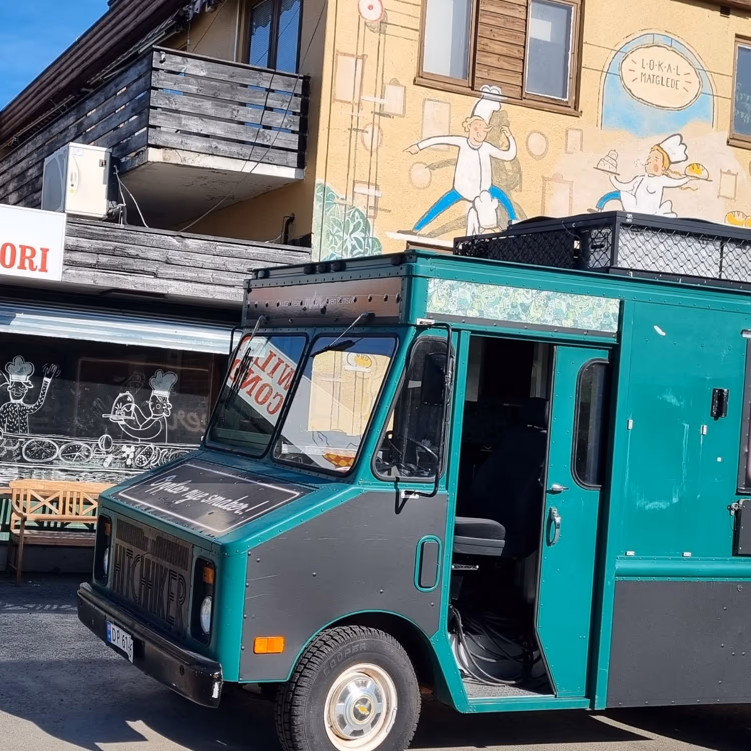 En grønn food truck står parkert på en gate foran en bygning med veggmalerier av kokker. Bygningen har en balkong og et restaurantskilt, og sollyset kaster tydelige skygger på scenen.