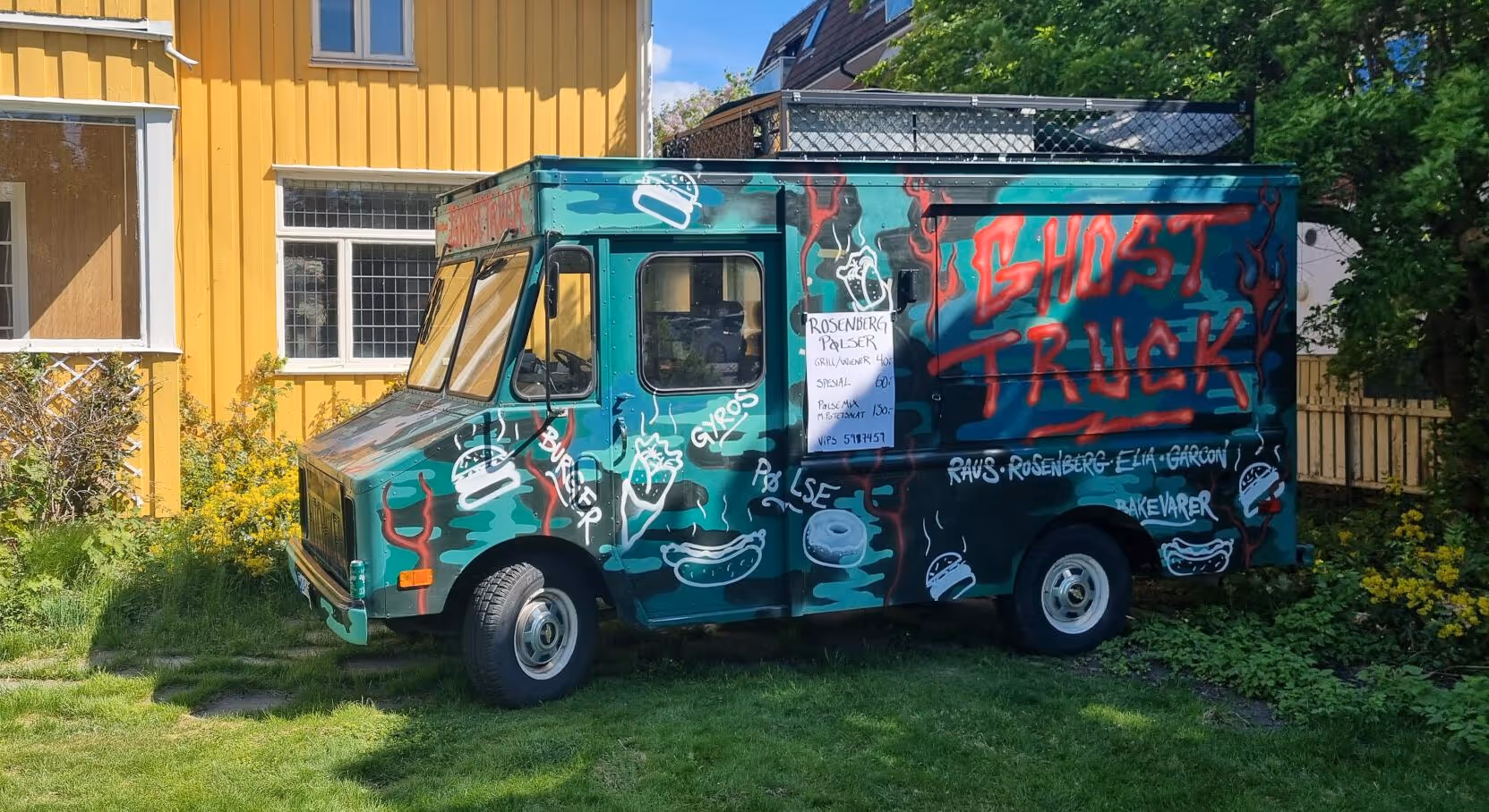 En fargerik foodtruck med graffitikunst, inkludert pølser og drikke, står parkert på gresset ved siden av et gult hus. "GHOST TRUCK" er sprayet med røde bokstaver på siden. Et skilt med en meny er teipet fast på lastebilen.
