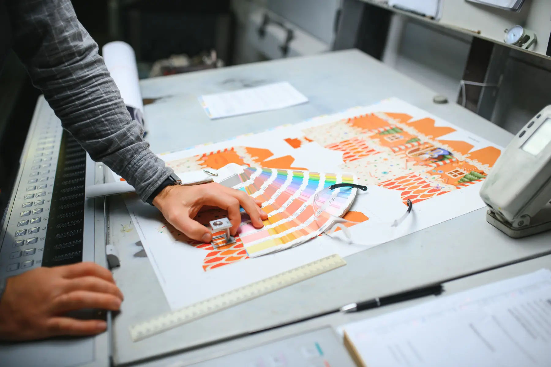A person inspecting printed color samples on a sheet using a magnifier on a worktable with safety glasses nearby.