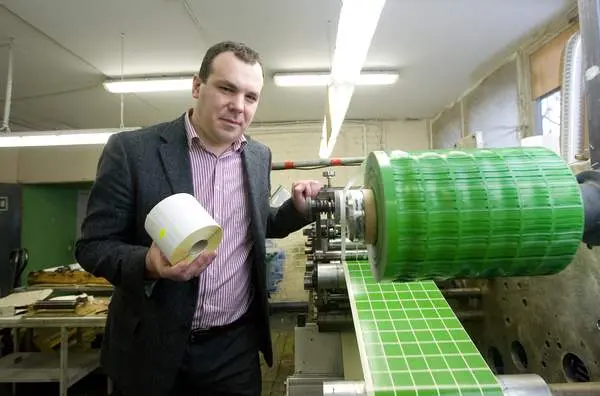 Man holding a roll of labels standing next to a machine printing green square labels indoors.