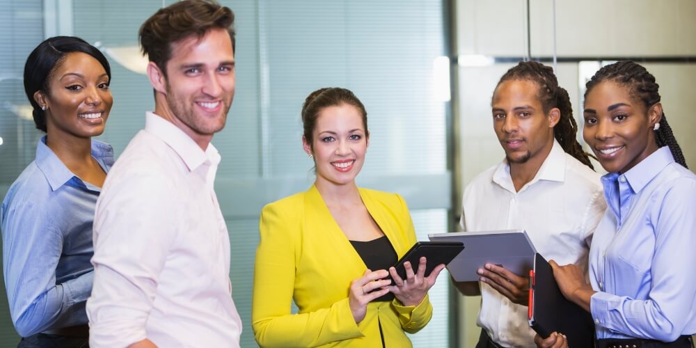 Five diverse business professionals standing together in an office, holding tablets and folders, engaged in a discussion.