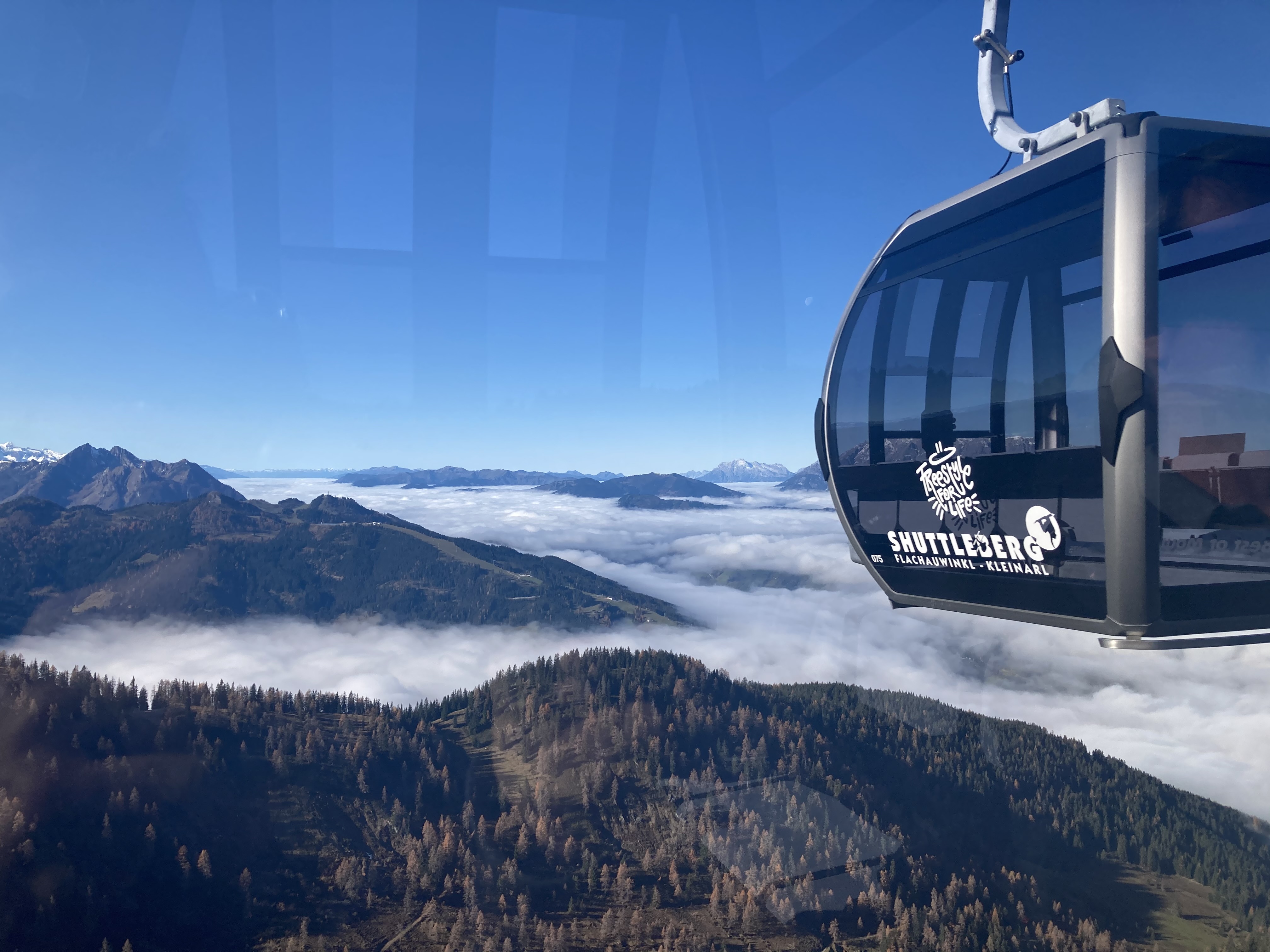 Gondel der Shuttleberg-Bahn schwebt über herbstlich bewaldeten Hängen mit Blick auf ein Nebelmeer und Bergpanorama