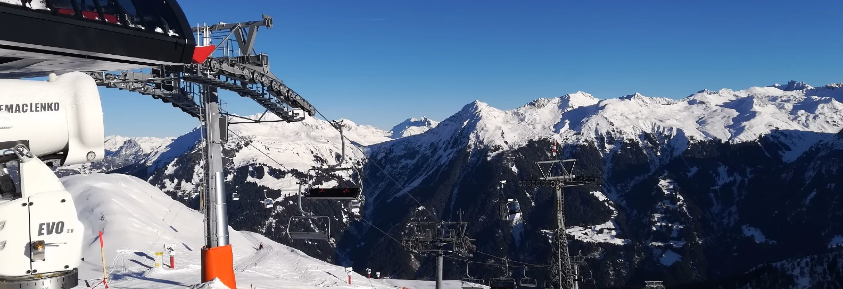 Sesselbahn mit Schneekanone vor beeindruckendem Alpenpanorama bei strahlend blauem Himmel