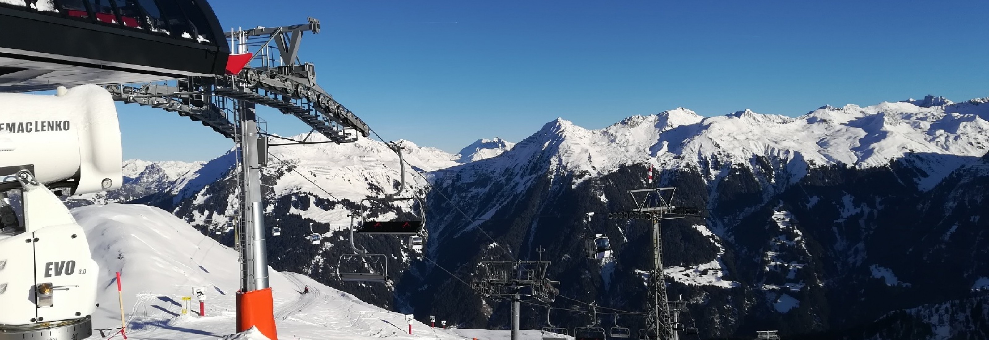 Sesselbahn mit Schneekanone vor beeindruckendem Alpenpanorama bei strahlend blauem Himmel