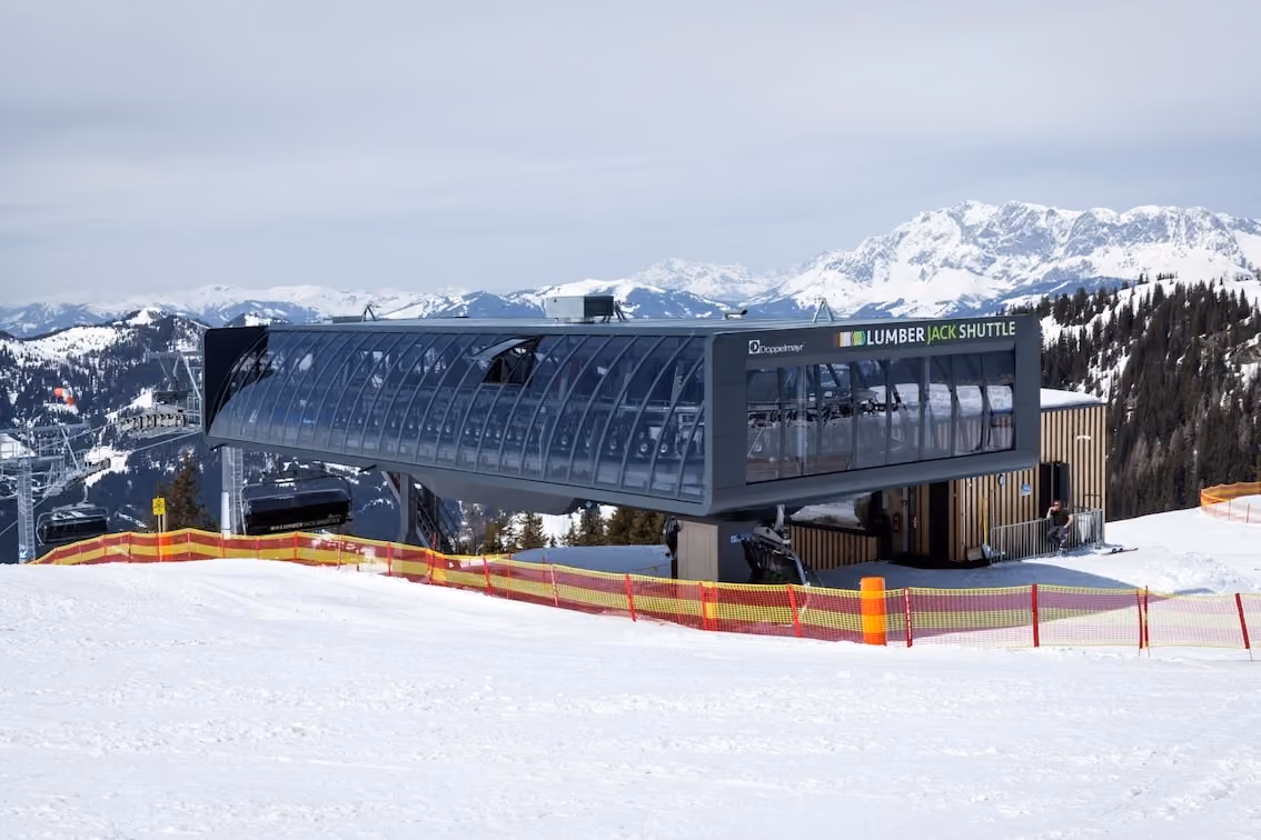 Moderne Sesselbahnstation „Lumberjack Shuttle“ vor verschneitem Panorama im Skigebiet mit umliegenden Bergen