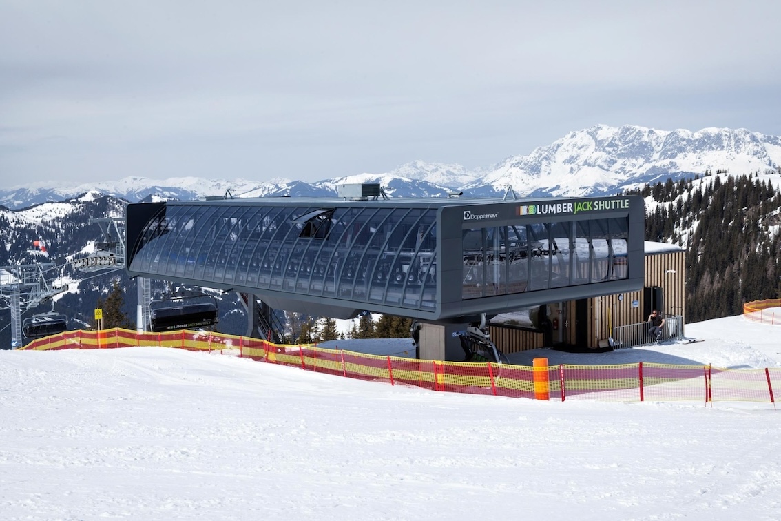 Moderne Sesselbahnstation „Lumberjack Shuttle“ vor verschneitem Panorama im Skigebiet mit umliegenden Bergen