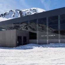 Talstation mit großflächiger Glasfassade und Betonbau vor schneebedecktem Bergpanorama bei klarem Himmel