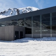 Talstation mit großflächiger Glasfassade und Betonbau vor schneebedecktem Bergpanorama bei klarem Himmel