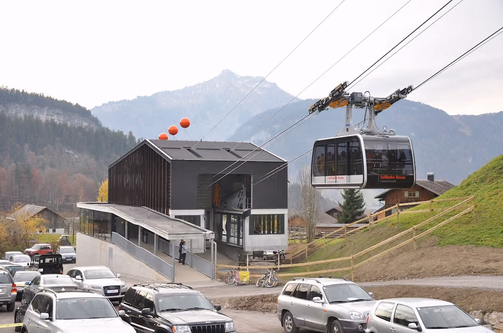 Talstation der Seilbahn Bezau mit ausfahrender Gondel und parkenden Autos im Vordergrund