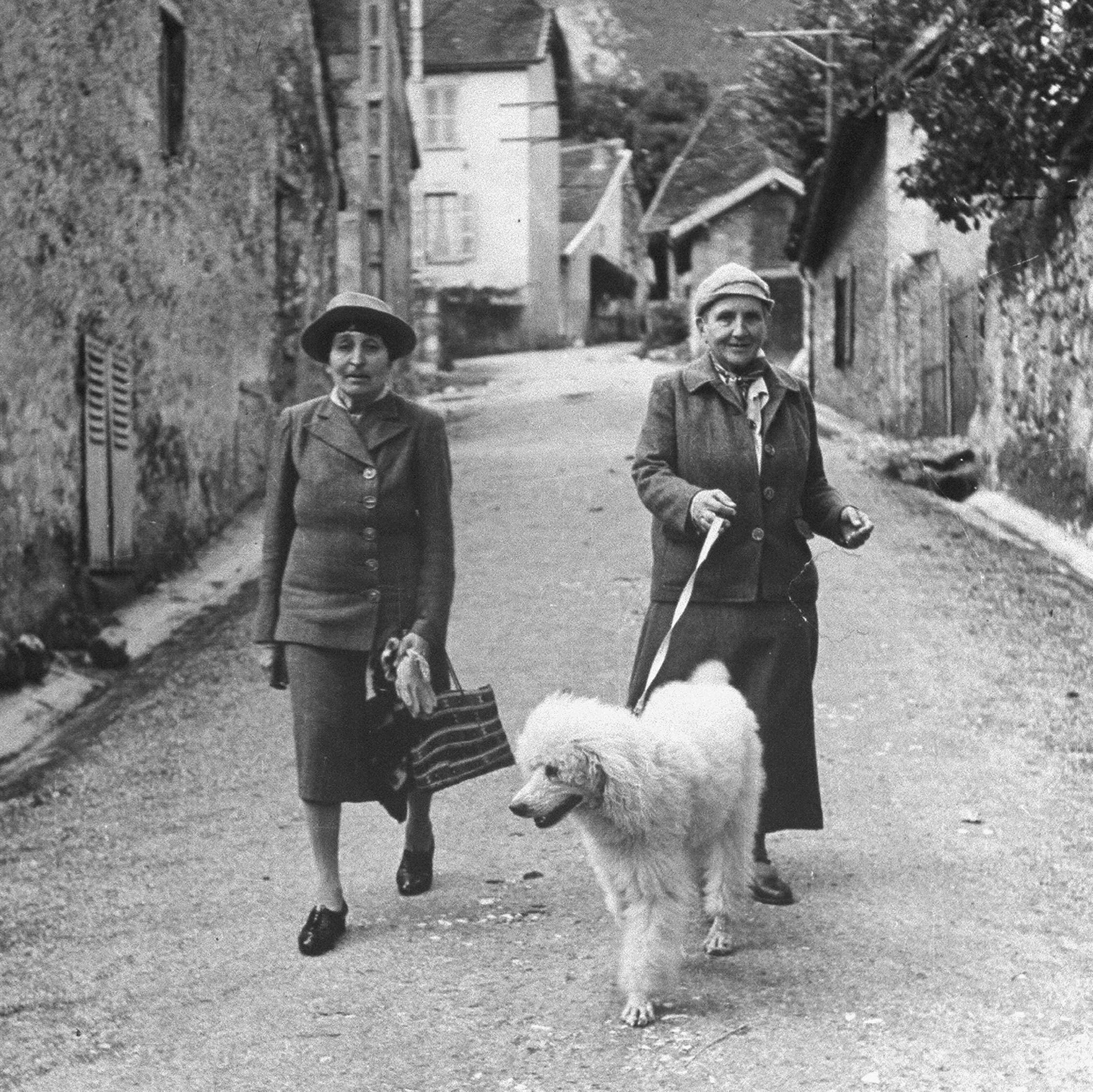 Black and white photo of Gertrude Stein & Alice B. Toklas walking their dag down a laneway.