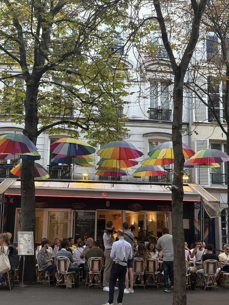 Front facade of the Le Ju bar in Paris, with patio and rainbow umbrellas hanging above.