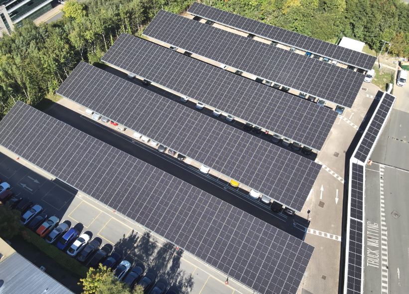 Aerial view of a solar carport structure with multiple rows of parked cars beneath large solar panel canopies in a parking lot surrounded by trees