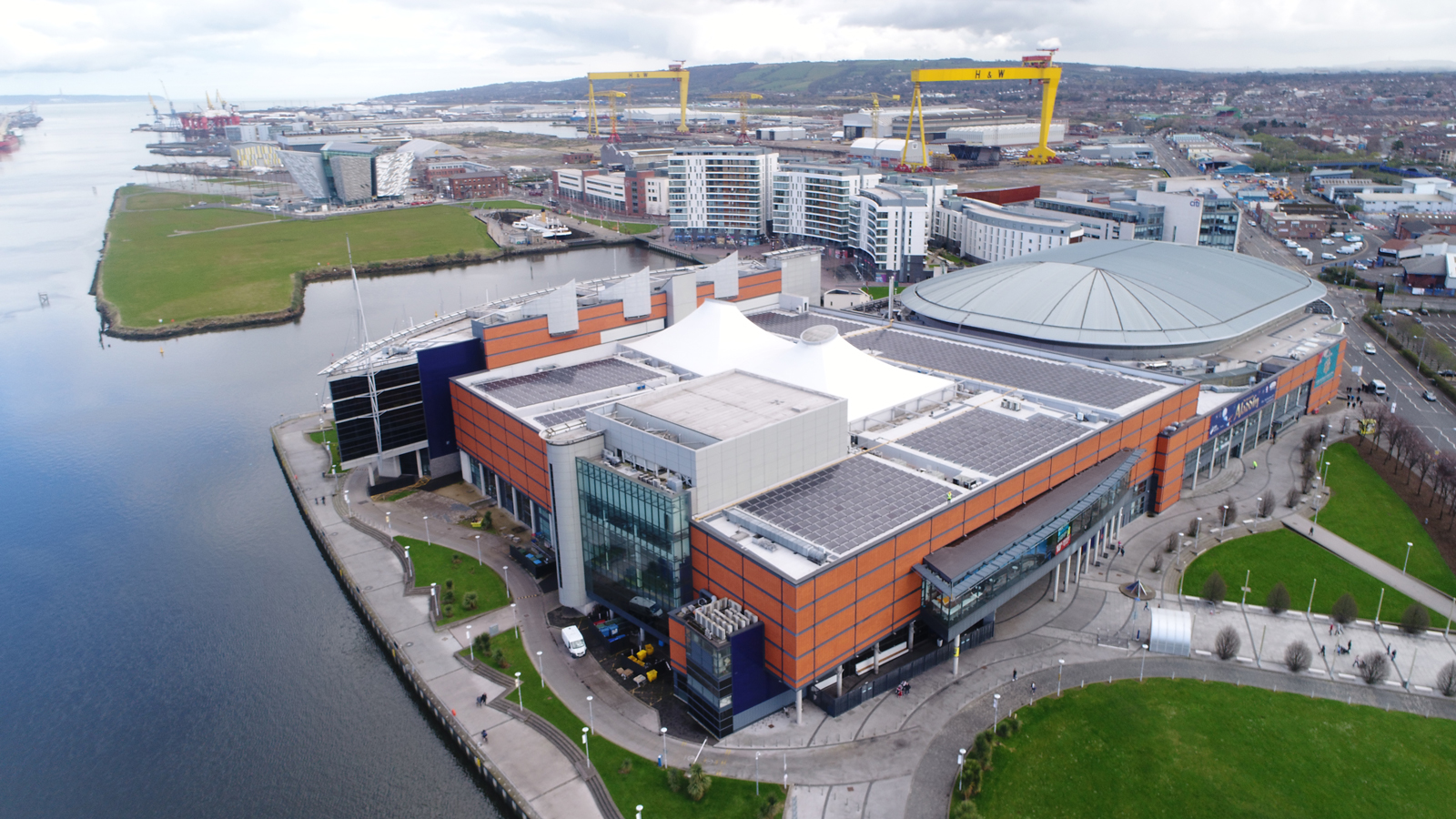Aerial view of SSE Arena Belfast with solar panels installed on the rooftop, located by the waterfront with Harland & Wolff cranes in the background