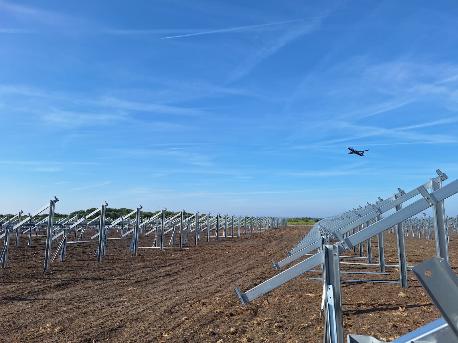 Early-stage construction of a solar farm at Liverpool John Lennon Airport with mounting frames installed and a plane flying overhead under a clear blue sky