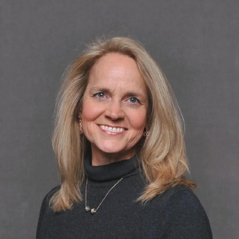 A woman with blonde hair smiling warmly in a dark turtleneck sweater and delicate necklace against a gray background.