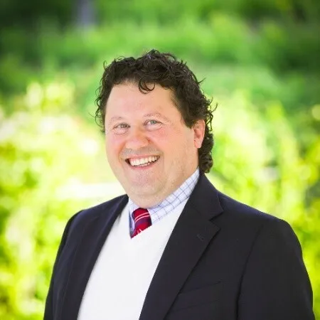 A man with curly dark hair smiling in a navy blazer and red tie, photographed outdoors against green foliage.